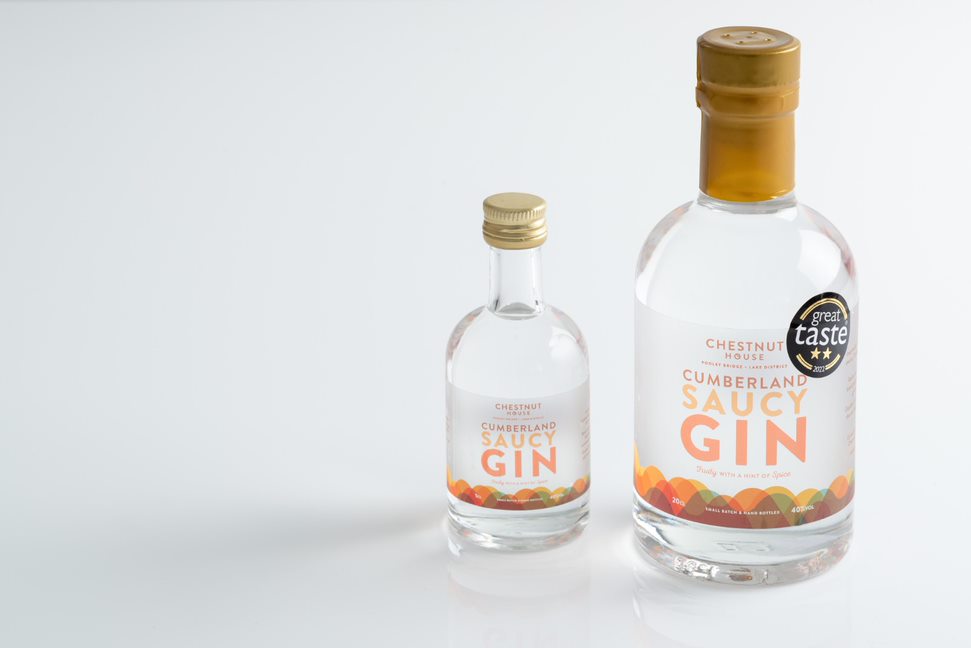 Two clear glass bottles of Chestnut House Cumberland Saucy Gin, one miniature and the other large, both featuring an orange lid.