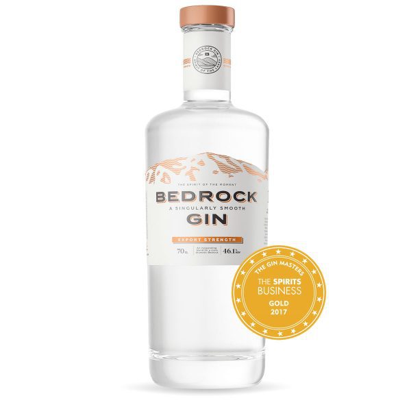 A clear glass bottle of Bedrock Export Gin. The bottle features a white and copper label with a mountain scene and a copper lid.