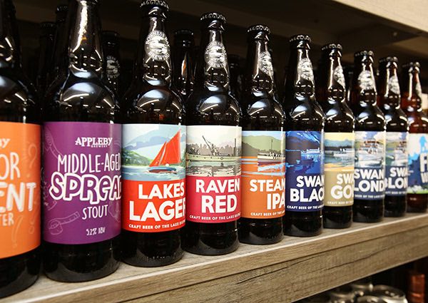 A shelf showcasing a row of dark glass bottles of Lake Districts craft beer from various brands with colourful labels